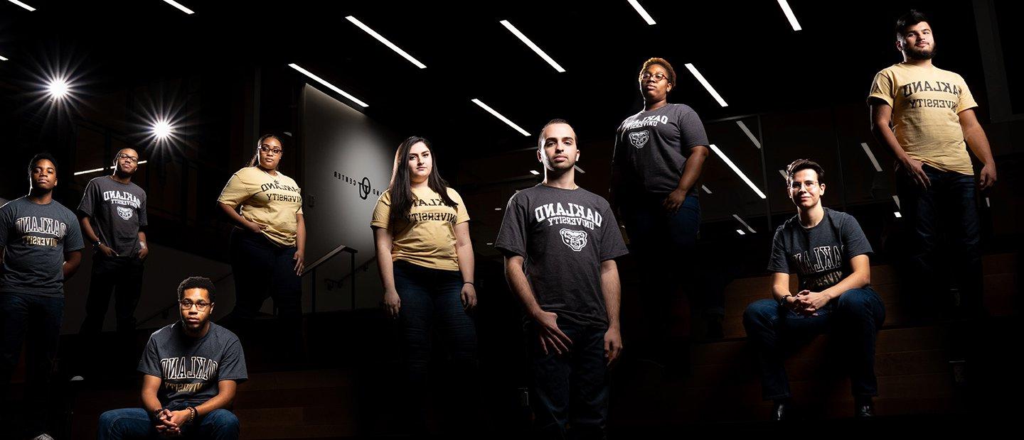 An artistic photo of a group of students, some standing and some sitting, in a dark room with spotlights.